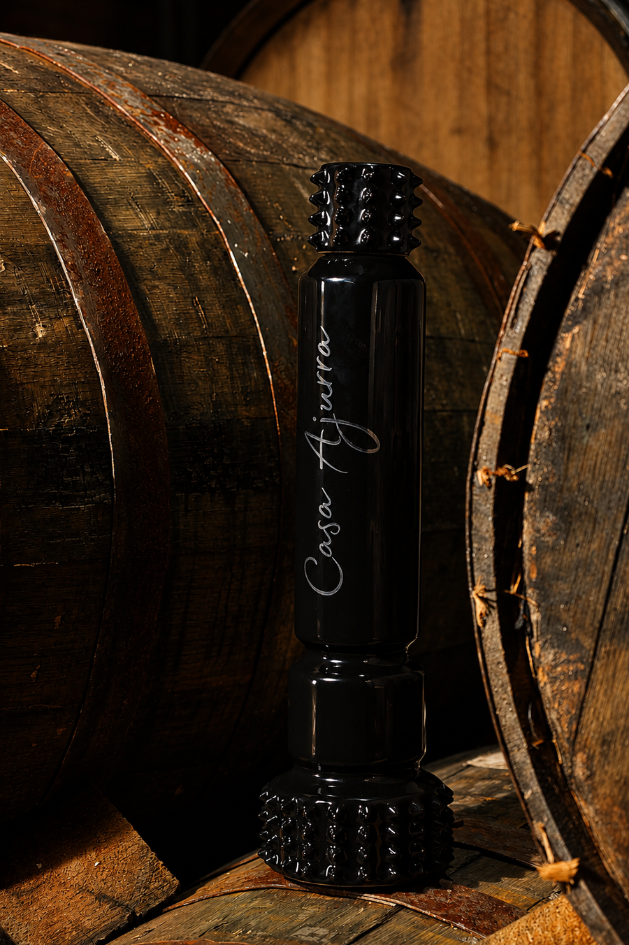 A black glass bottle with a textured screw cap and base, featuring the white script logo 'Casa Agüera', set against a rustic background of wooden wine barrels.