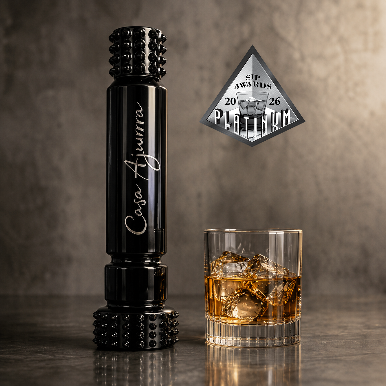 A black whiskey glass stopper with silver script engraving reading 'Casa Aijurra' stands on a dark surface beside a rocks glass filled with amber whiskey and ice. A silver award badge reading '50 Awards 2026 Platinum' floats above against a textured gray background.